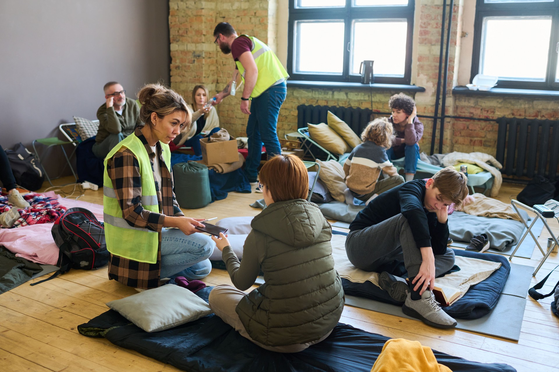 Young female volunteer passing smartphone to one of refugees sitting on mattress